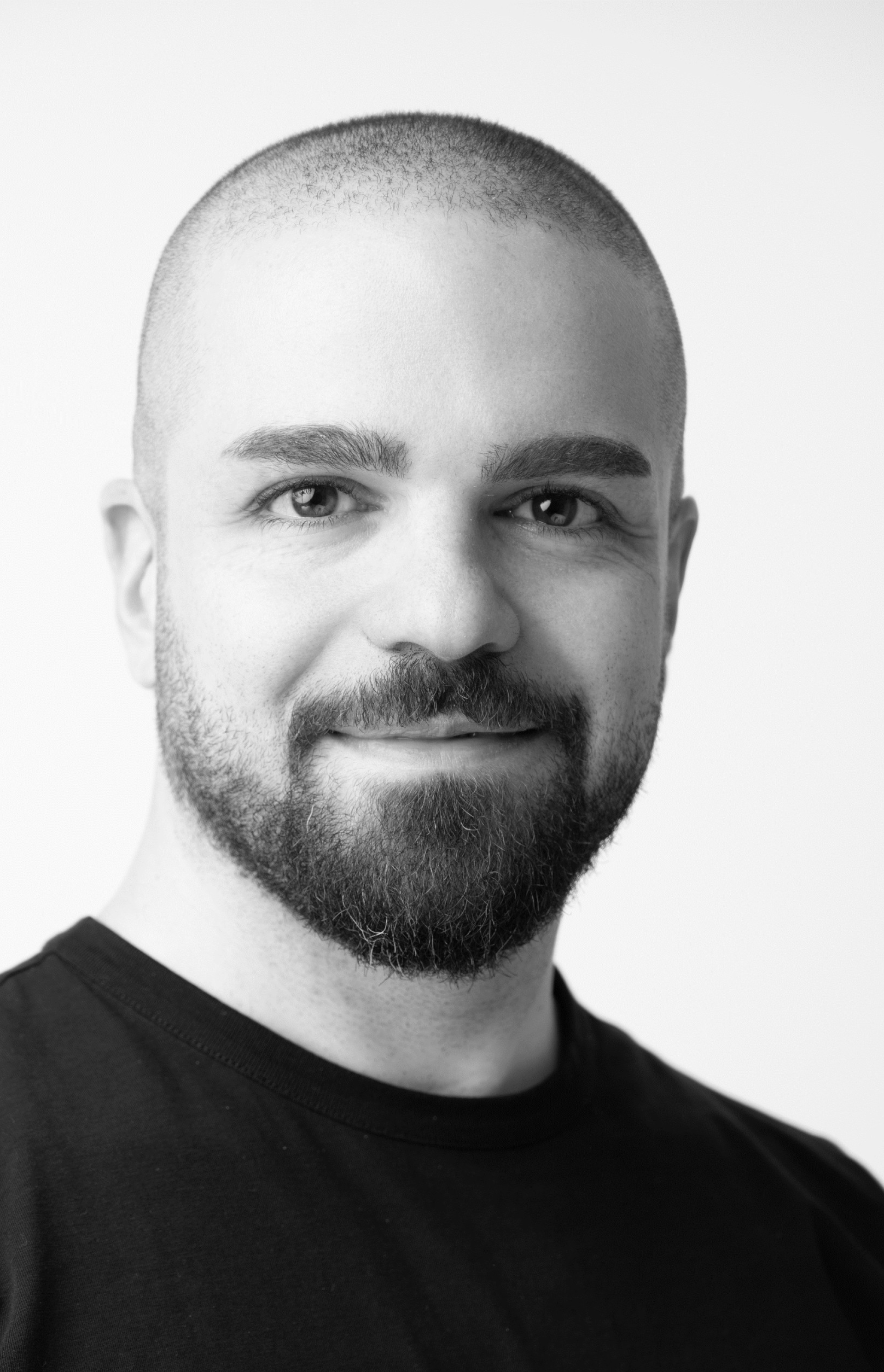 Black-and-white headshot of a person with a shaved head and short beard, wearing a dark crewneck shirt, facing forward against a plain light background.